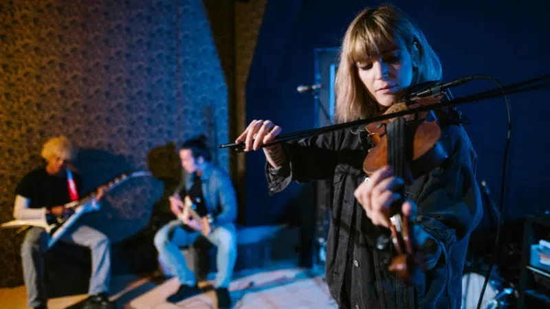 Una mujer toca el violín mientras otros dos músicos tocan guitarras en el fondo, todos interpretando en un espacio interior tenuemente iluminado—mostrando cómo construir un negocio musical escalable a través de presentaciones en vivo cautivadoras.
