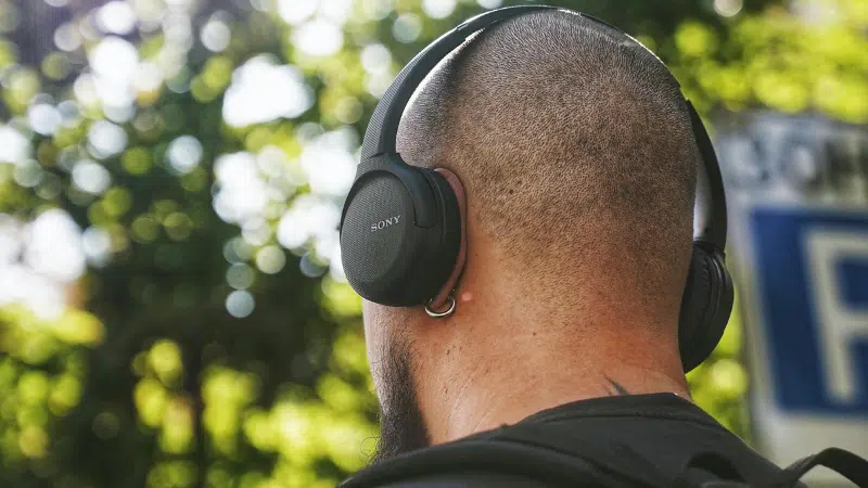 Una persona con la cabeza rapada y una mochila revisa su email mientras usa audífonos negros Sony over-ear al aire libre, con árboles de fondo.