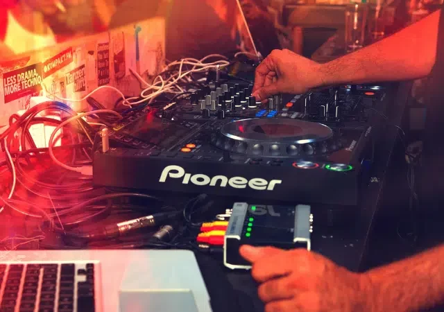 Una persona maneja un controlador Pioneer DJ con varios cables conectados, junto a una laptop con Spotify abierto, en un ambiente tenue iluminado con luces de colores.