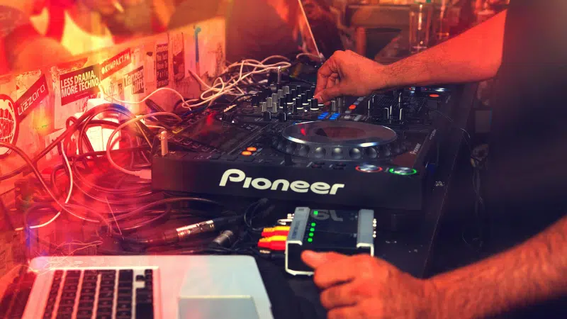 Una persona maneja un controlador Pioneer DJ con varios cables conectados, junto a una laptop con Spotify abierto, en un ambiente tenue iluminado con luces de colores.
