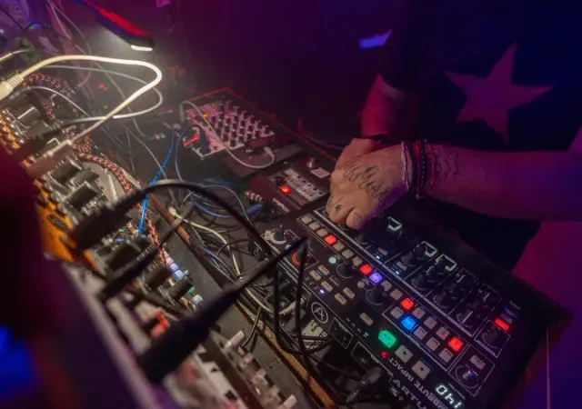 Un distribuidor opera con destreza una variedad de equipos de música electrónica y sintetizadores, iluminado por luces de escenario coloridas.