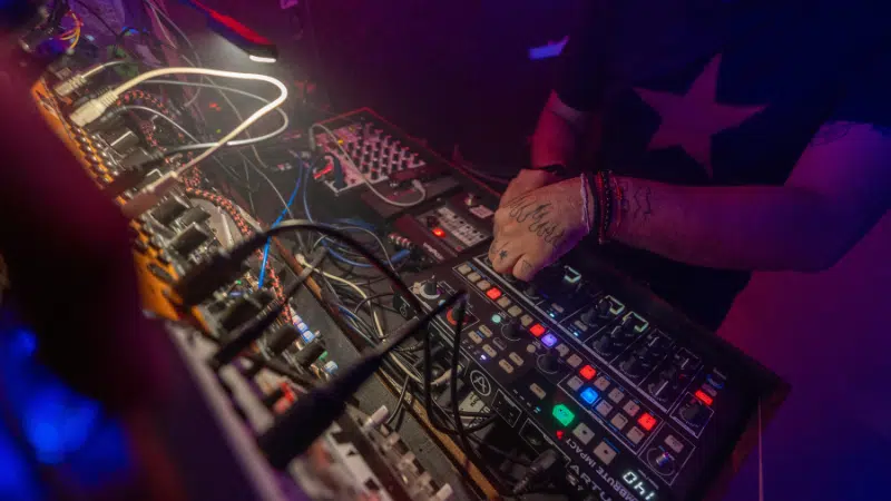 Un distribuidor opera con destreza una variedad de equipos de música electrónica y sintetizadores, iluminado por luces de escenario coloridas.