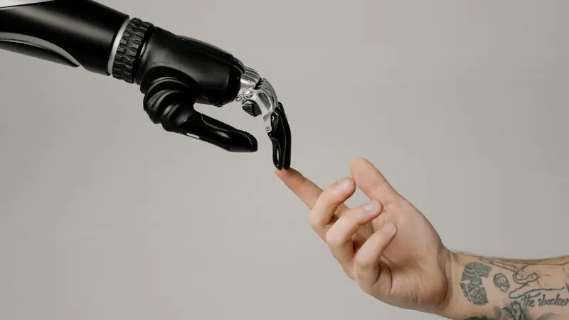 Una mano robótica y una mano humana se extienden una hacia la otra, con los dedos casi tocándose sobre un fondo sencillo, simbolizando la intersección entre tecnología y humanidad en una escena que recuerda a imágenes icónicas del copyright.