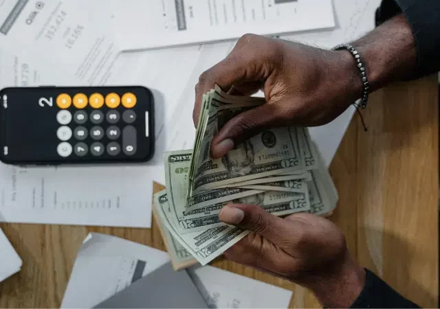 Una persona cuenta billetes de dólares en un escritorio con una calculadora, documentos financieros esparcidos y la aplicación SplitShare abierta en su pantalla.