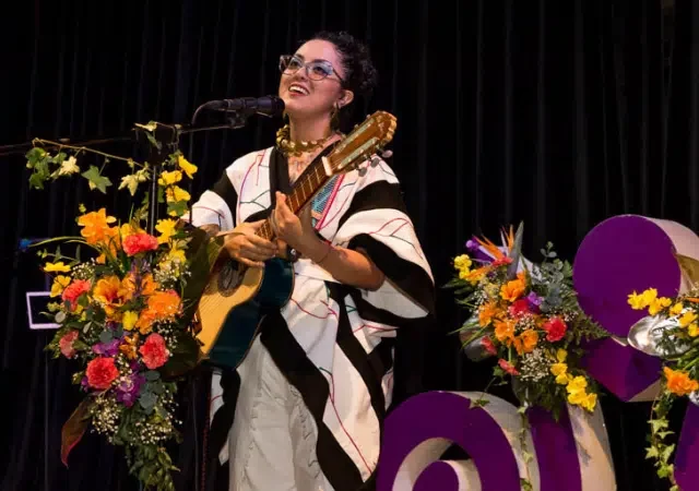 Una mujer con un atuendo tradicional latinoamericano toca una guitarra y canta en un micrófono en el escenario, rodeada de arreglos florales coloridos y decoraciones moradas.