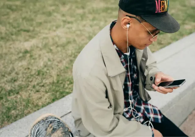 Una persona sentada en un borde de concreto al aire libre, usando una gorra, gafas, audífonos y una chaqueta beige, mirando su teléfono inteligente—quizás revisando Spotify. Se ve césped en el fondo.