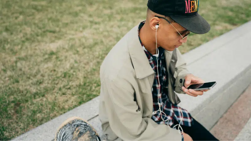 Una persona sentada en un borde de concreto al aire libre, usando una gorra, gafas, audífonos y una chaqueta beige, mirando su teléfono inteligente—quizás revisando Spotify. Se ve césped en el fondo.
