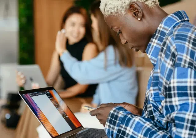 Una persona con una camisa de cuadros usa un smartphone y una laptop, posiblemente revisando Amazon Music Insights, mientras otras dos personas con laptops conversan en el fondo.