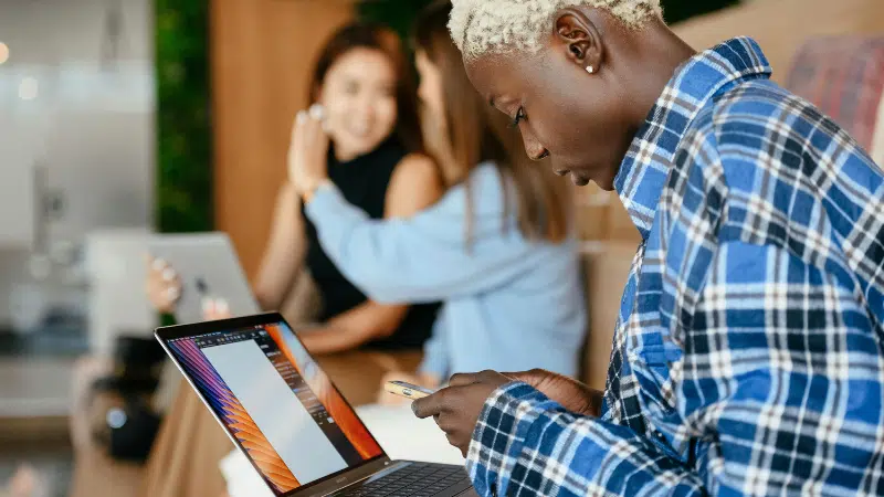 Una persona con una camisa de cuadros usa un smartphone y una laptop, posiblemente revisando Amazon Music Insights, mientras otras dos personas con laptops conversan en el fondo.