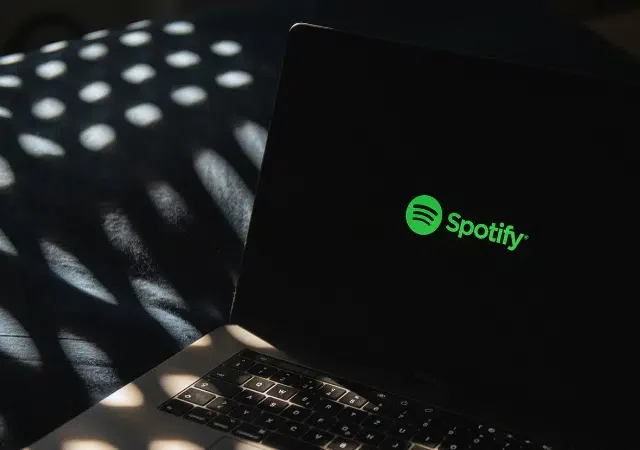 Un portátil con el logo de Spotify en la pantalla se encuentra sobre una superficie oscura, rodeado de patrones de luz moteada que le dan un toque atmosférico.