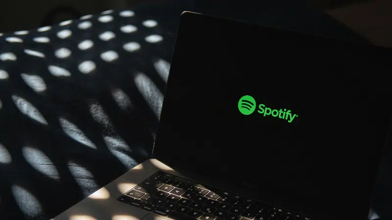Un portátil con el logo de Spotify en la pantalla se encuentra sobre una superficie oscura, rodeado de patrones de luz moteada que le dan un toque atmosférico.