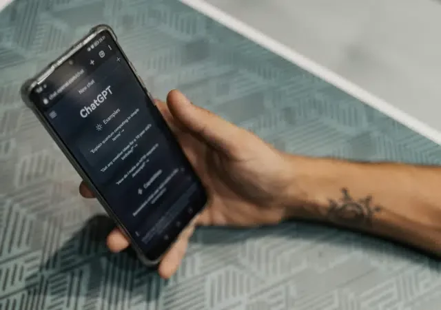 Una persona sostiene un smartphone que muestra el sitio web de ChatGPT sobre una mesa con patrones geométricos, ilustrando cómo ChatGPT se integra perfectamente en la vida cotidiana.