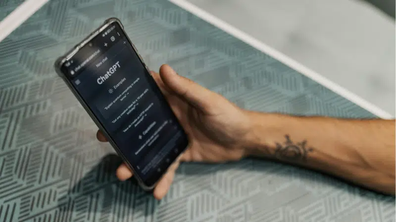Una persona sostiene un smartphone que muestra el sitio web de ChatGPT sobre una mesa con patrones geométricos, ilustrando cómo ChatGPT se integra perfectamente en la vida cotidiana.