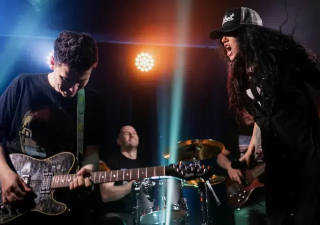 Una banda de rock se presenta en el escenario con un guitarrista, baterista, bajista y un vocalista con sombrero que canta apasionadamente bajo luces de colores, atrayendo a la multitud hacia su energético embudo de fans.