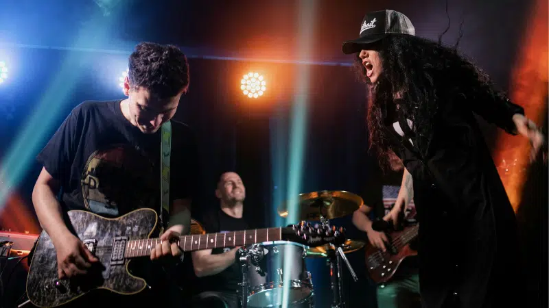 Una banda de rock se presenta en el escenario con un guitarrista, baterista, bajista y un vocalista con sombrero que canta apasionadamente bajo luces de colores, atrayendo a la multitud hacia su energético embudo de fans.