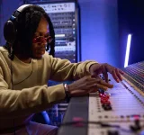 Una persona con audífonos y gafas de sol ajusta los controles de una gran consola de audio, gestionando contenido en un estudio profesional.