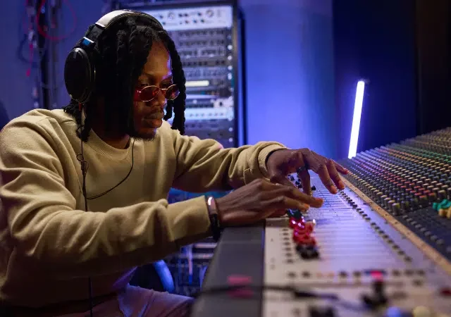 Una persona con audífonos y gafas de sol ajusta los controles de una gran consola de audio, gestionando contenido en un estudio profesional.