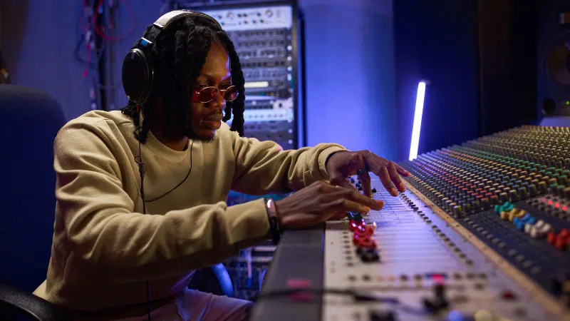 Una persona con audífonos y gafas de sol ajusta los controles de una gran consola de audio, gestionando contenido en un estudio profesional.