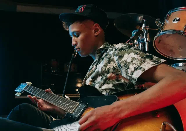 Un joven con camisa floral y gorra toca una guitarra eléctrica en el escenario, con una batería y luces detrás, celebrando el lanzamiento de nueva música.