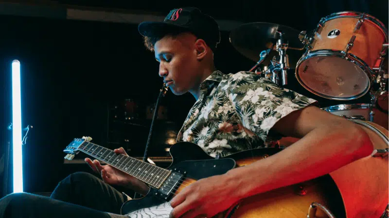 Un joven con camisa floral y gorra toca una guitarra eléctrica en el escenario, con una batería y luces detrás, celebrando el lanzamiento de nueva música.