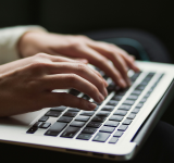 Primer plano de una persona escribiendo en el teclado de una laptop, con sus manos moviéndose en un ritmo sinfónico, mientras la pantalla es parcialmente visible al fondo.