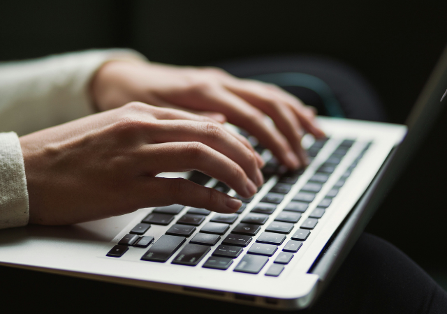 Primer plano de una persona escribiendo en el teclado de una laptop, con sus manos moviéndose en un ritmo sinfónico, mientras la pantalla es parcialmente visible al fondo.