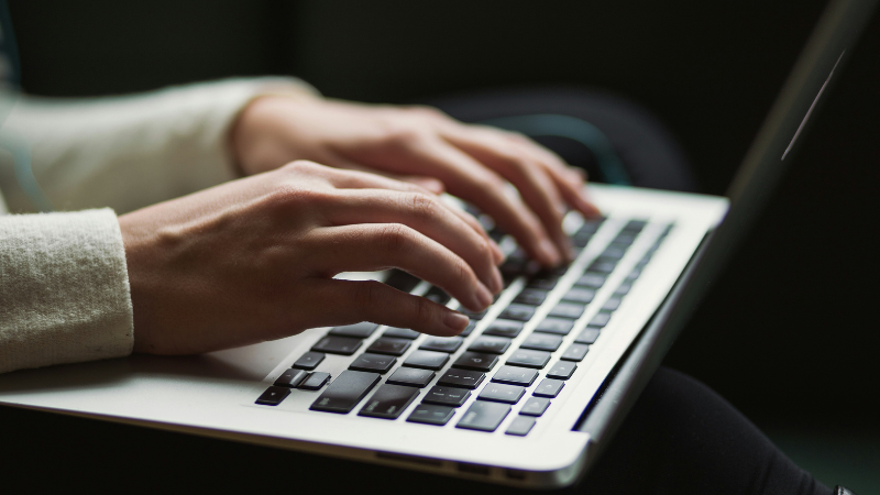 Primer plano de una persona escribiendo en el teclado de una laptop, con sus manos moviéndose en un ritmo sinfónico, mientras la pantalla es parcialmente visible al fondo.