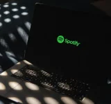 Una laptop que muestra el logo verde de Spotify en la pantalla descansa sobre una cama iluminada por la luz del sol, insinuando un espacio acogedor donde la música y la tecnología de IA se encuentran.