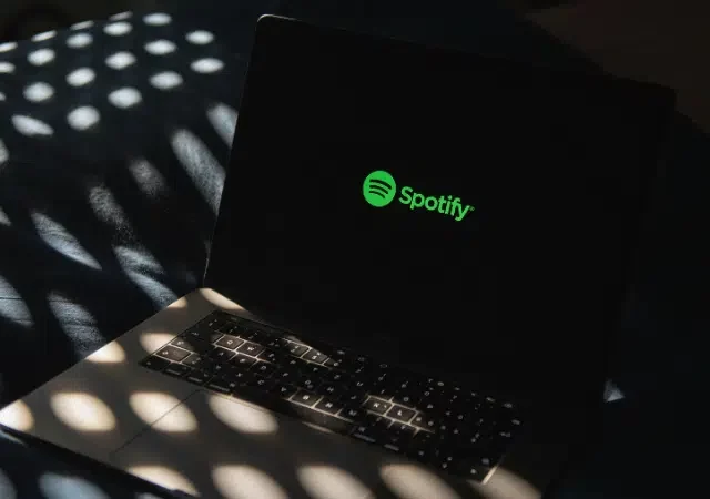 Una laptop que muestra el logo verde de Spotify en la pantalla descansa sobre una cama iluminada por la luz del sol, insinuando un espacio acogedor donde la música y la tecnología de IA se encuentran.