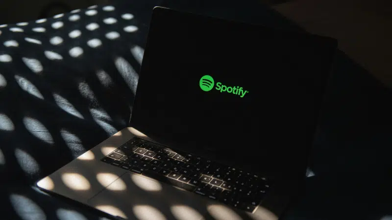 Una laptop que muestra el logo verde de Spotify en la pantalla descansa sobre una cama iluminada por la luz del sol, insinuando un espacio acogedor donde la música y la tecnología de IA se encuentran.