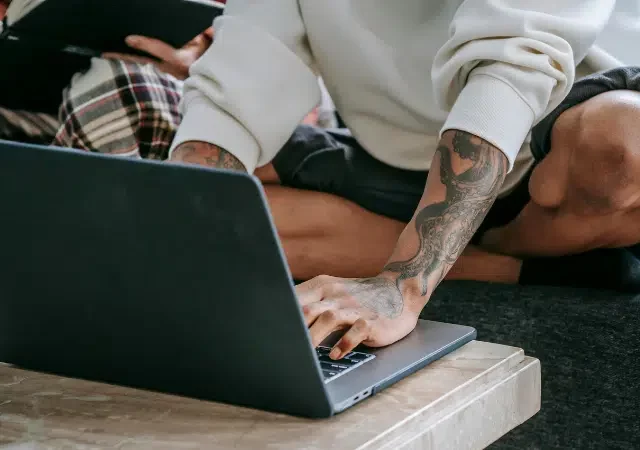 Una persona con tatuajes está sentada con las piernas cruzadas en un sofá usando herramientas digitales—una laptop sobre la mesa—mientras otra persona al fondo sostiene una tablet.