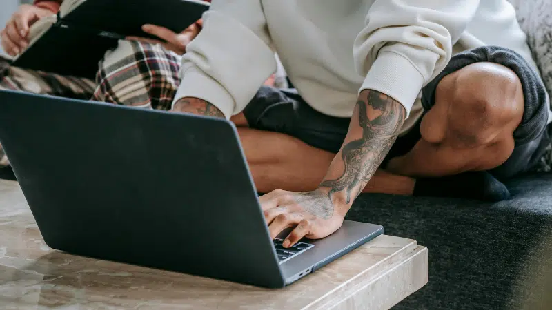 Una persona con tatuajes está sentada con las piernas cruzadas en un sofá usando herramientas digitales—una laptop sobre la mesa—mientras otra persona al fondo sostiene una tablet.