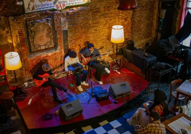 Tres músicos tocan guitarras y teclado en un pequeño escenario de un acogedor local en Bogotá, con paredes de ladrillo rojo y decoración vintage.