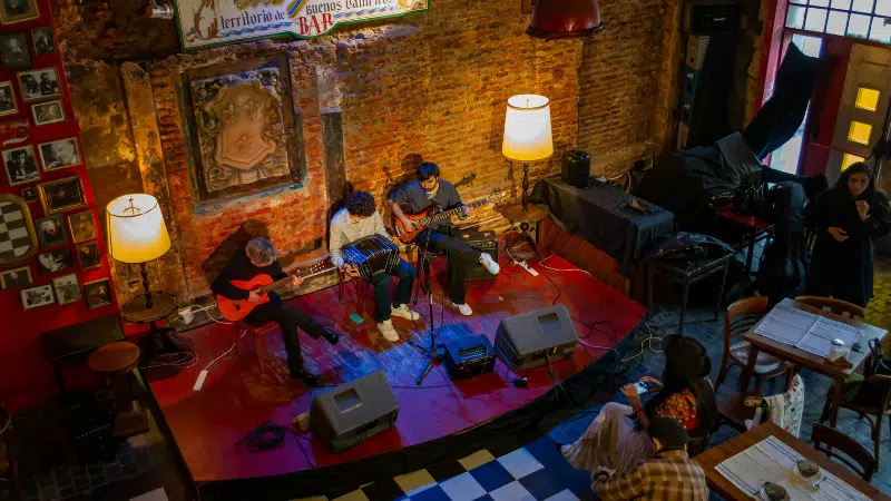 Tres músicos tocan guitarras y teclado en un pequeño escenario de un acogedor local en Bogotá, con paredes de ladrillo rojo y decoración vintage.
