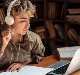 Una persona con audífonos estudia en un escritorio, inmersa en melodías sinfónicas, rodeada de una laptop, un cuaderno, un libro abierto y una taza de café en una biblioteca o sala de estudio tranquila.