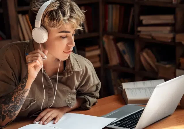 Una persona con audífonos estudia en un escritorio, inmersa en melodías sinfónicas, rodeada de una laptop, un cuaderno, un libro abierto y una taza de café en una biblioteca o sala de estudio tranquila.