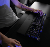 Una persona usando un teclado mecánico y un mouse sobre un escritorio negro; solo se ven sus brazos y torso.