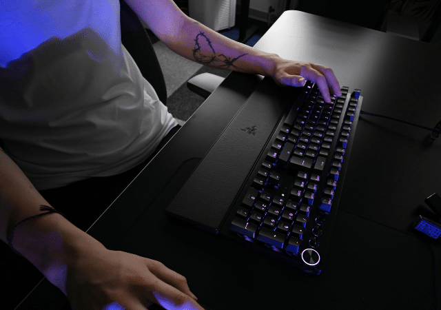 Una persona usando un teclado mecánico y un mouse sobre un escritorio negro; solo se ven sus brazos y torso.