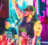 Una artista en el escenario levanta un brazo mientras canta en un micrófono, capturando la emoción de empezar, con luces de colores brillando y el público levantando las manos en primer plano.