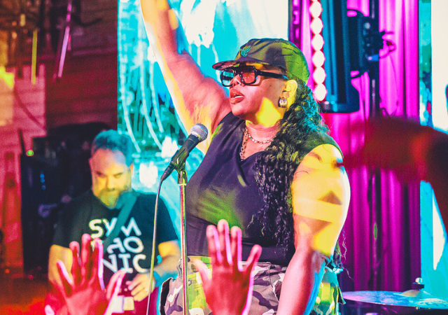 Una artista en el escenario levanta un brazo mientras canta en un micrófono, capturando la emoción de empezar, con luces de colores brillando y el público levantando las manos en primer plano.