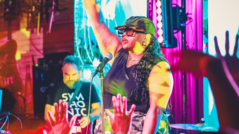 Una artista en el escenario levanta un brazo mientras canta en un micrófono, capturando la emoción de empezar, con luces de colores brillando y el público levantando las manos en primer plano.