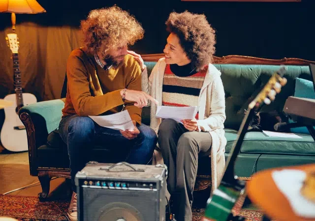 Dos personas estan sentadas en un sofa verde, sosteniendo papeles y hablando sobre DSPs entre instrumentos musicales, incluidas guitarras y un teclado, en una habitacion acogedora y con poca luz.