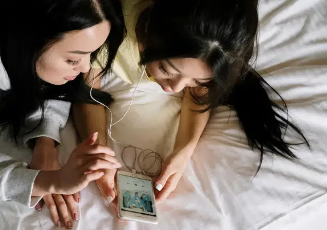 Dos personas acostadas en una cama comparten unos audífonos, disfrutando Apple Music mientras miran un smartphone juntas.