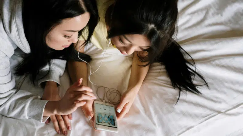 Dos personas acostadas en una cama comparten unos audífonos, disfrutando Apple Music mientras miran un smartphone juntas.