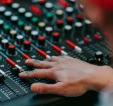 Una persona ajusta deslizadores y perillas en una consola profesional de mezcla de audio, con varios botones y controles de colores visibles.
