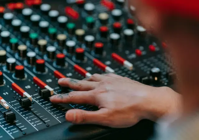 Una persona ajusta deslizadores y perillas en una consola profesional de mezcla de audio, con varios botones y controles de colores visibles.