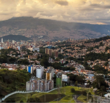 Una vista panorámica de Medellín con edificios densamente agrupados entre colinas verdes y montañas bajo un cielo nublado al atardecer.