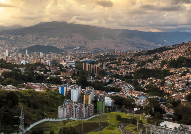 Una vista panorámica de Medellín con edificios densamente agrupados entre colinas verdes y montañas bajo un cielo nublado al atardecer.
