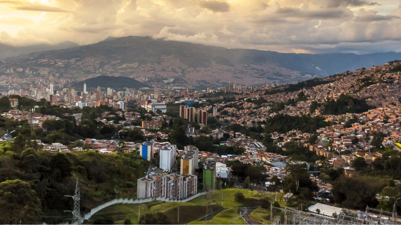 Una vista panorámica de Medellín con edificios densamente agrupados entre colinas verdes y montañas bajo un cielo nublado al atardecer.