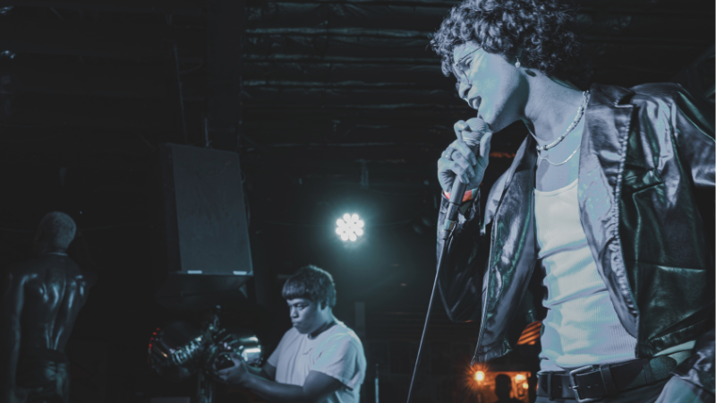 Un cantante con chaqueta de cuero actúa en el escenario con un micrófono, mientras un guitarrista toca en segundo plano bajo una iluminación azul, estrenando temas de su último lanzamiento.
